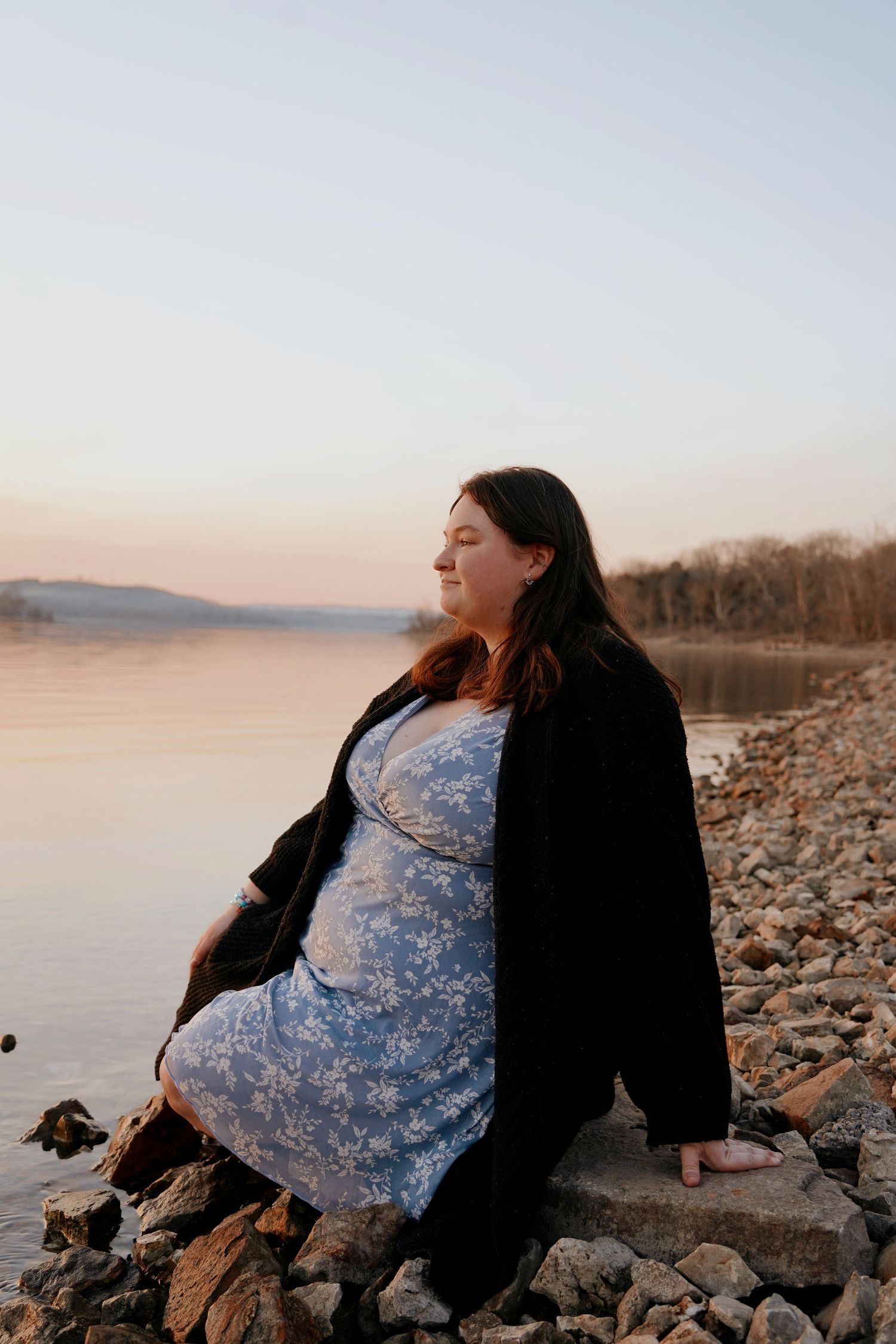 Plus-size woman in a floral dress and black cardigan sits on rocky shoreline at sunset.