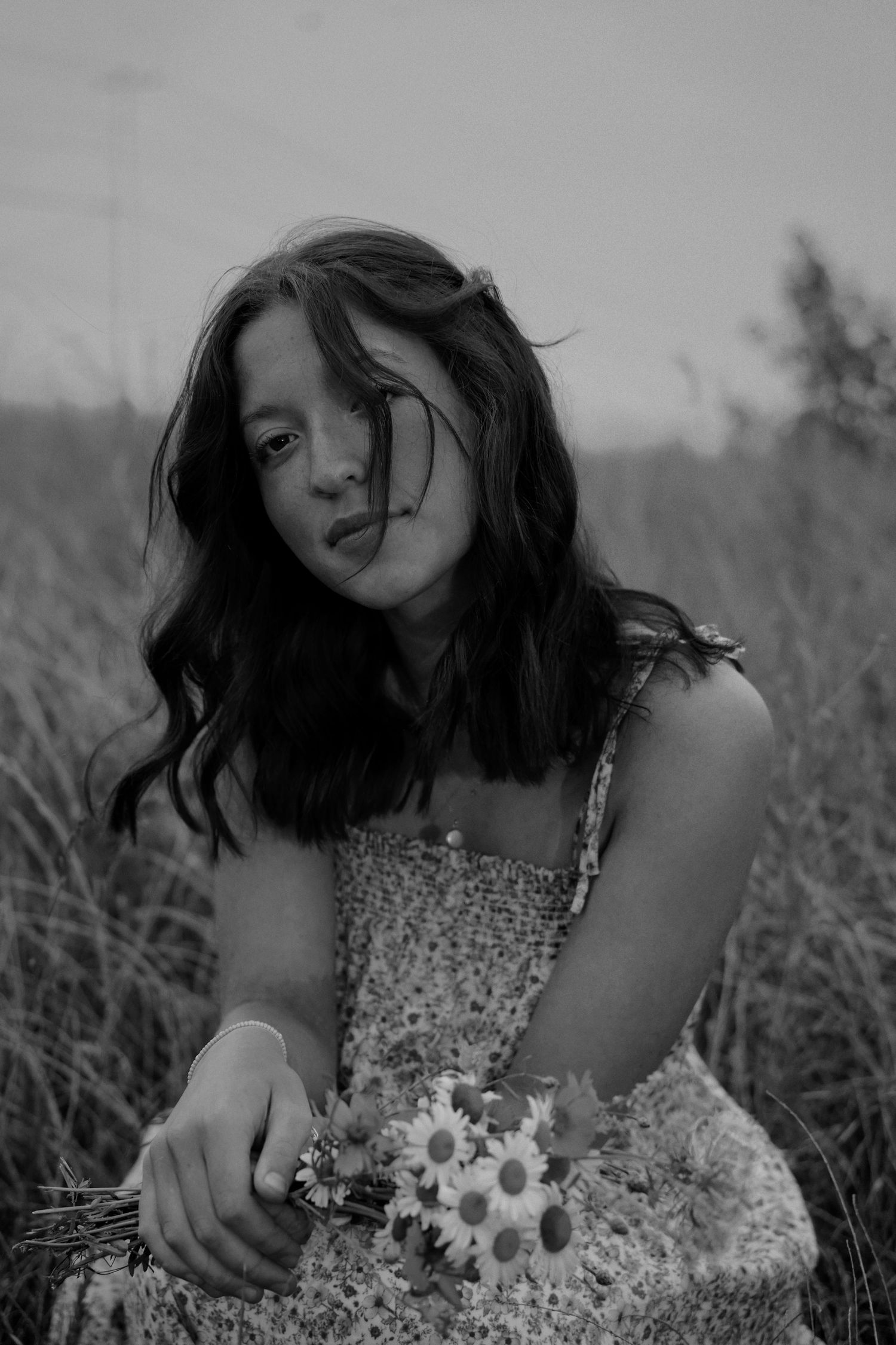 Black and white portrait of a young woman holding wildflowers in a windswept grassy field.