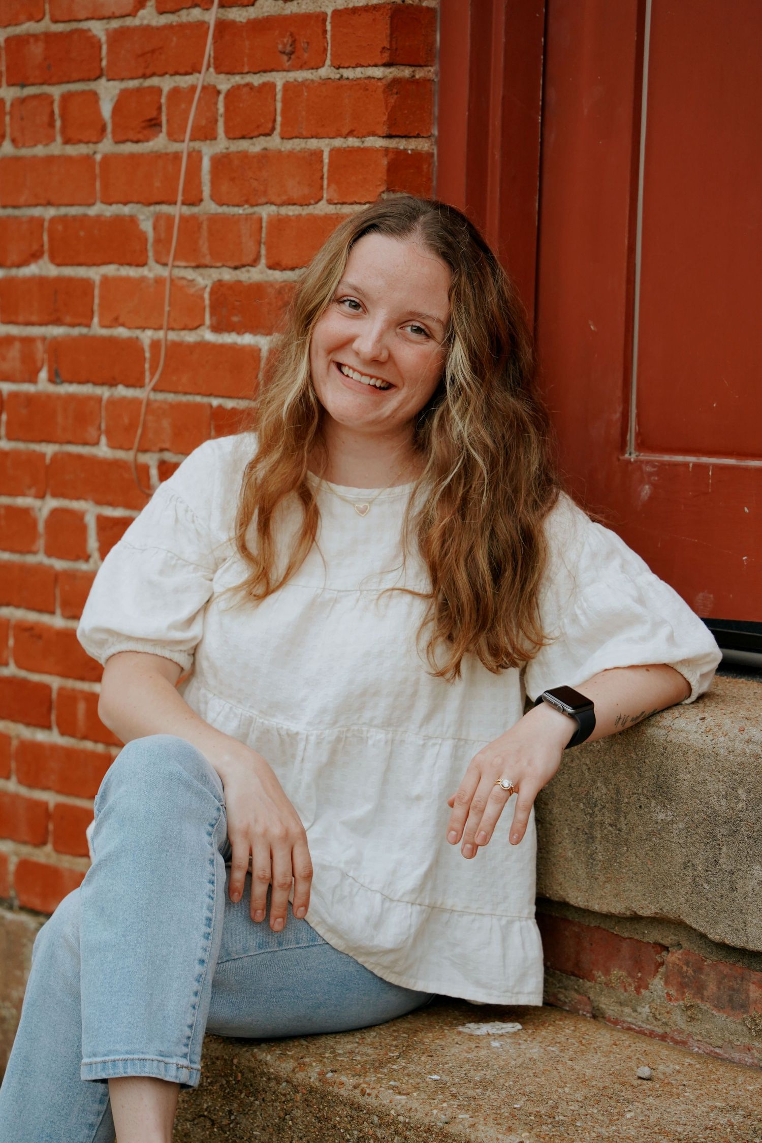 Smiling young woman in a white blouse and jeans sits on steps against a red brick wall.