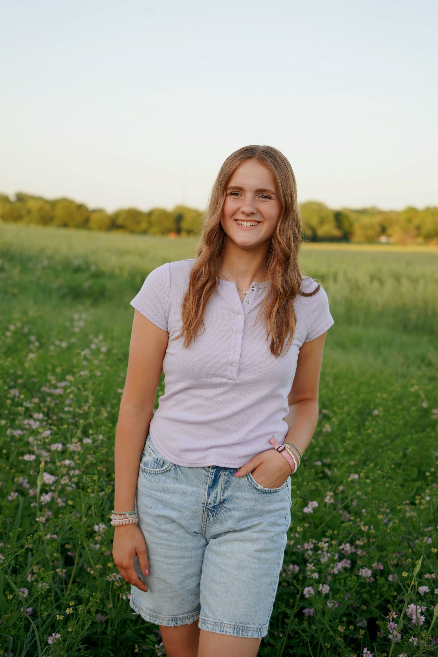 Smiling teenage girl in a pink top and denim shorts stands in a green flowering meadow at sunset.