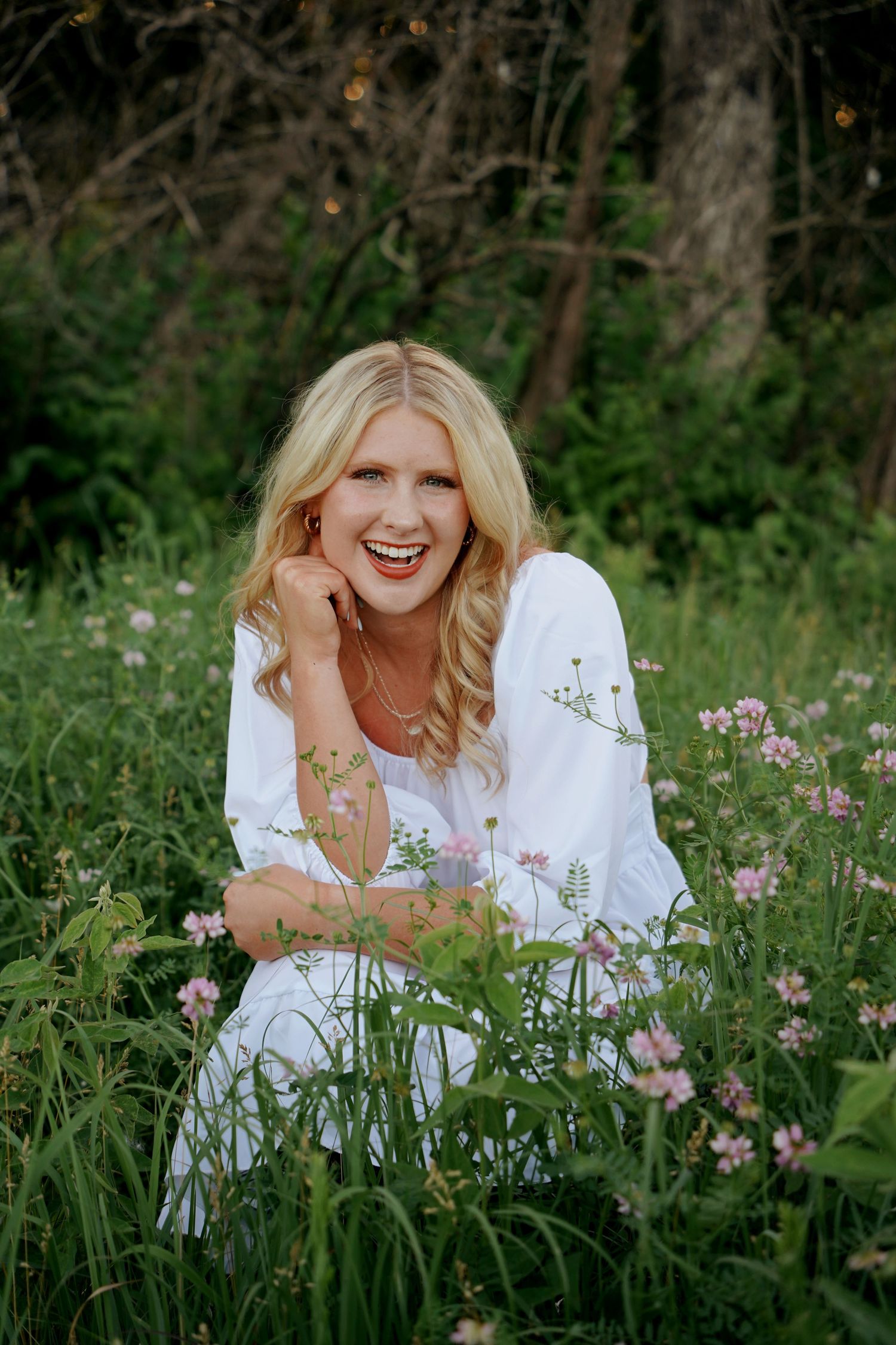 Laughing blonde woman in a white dress sits among pink wildflowers in a lush green forest clearing.