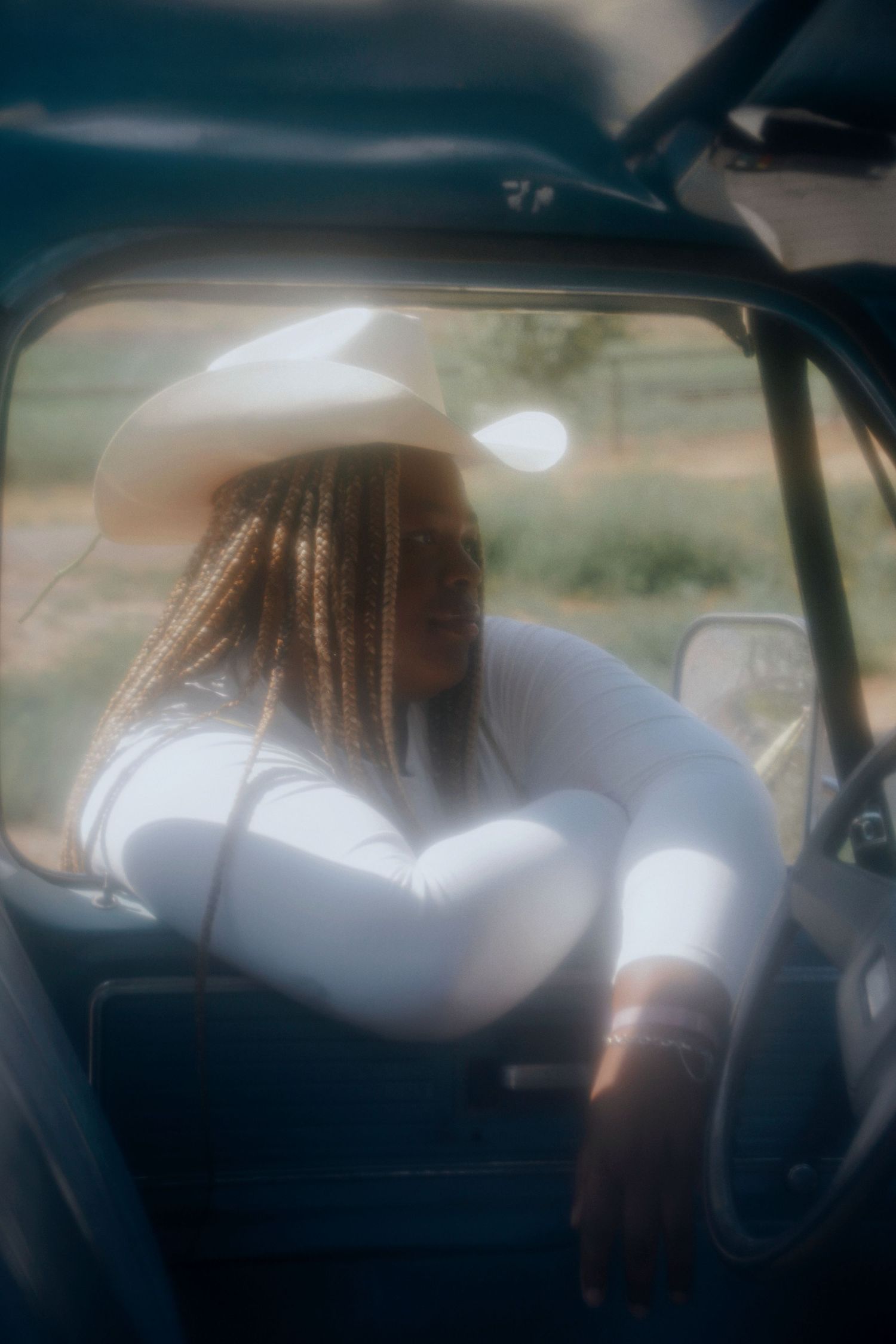 Young woman in a white cowboy hat and top leans out of a vintage truck window in soft light.