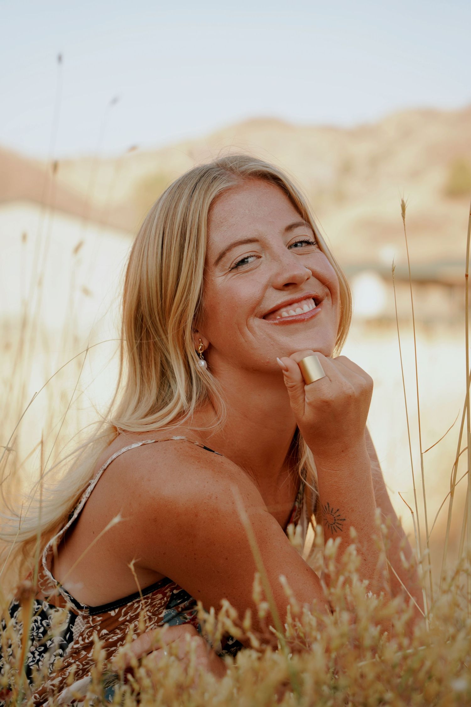 Smiling blonde woman with a gold ring poses among tall dry grass in warm golden sunlight.