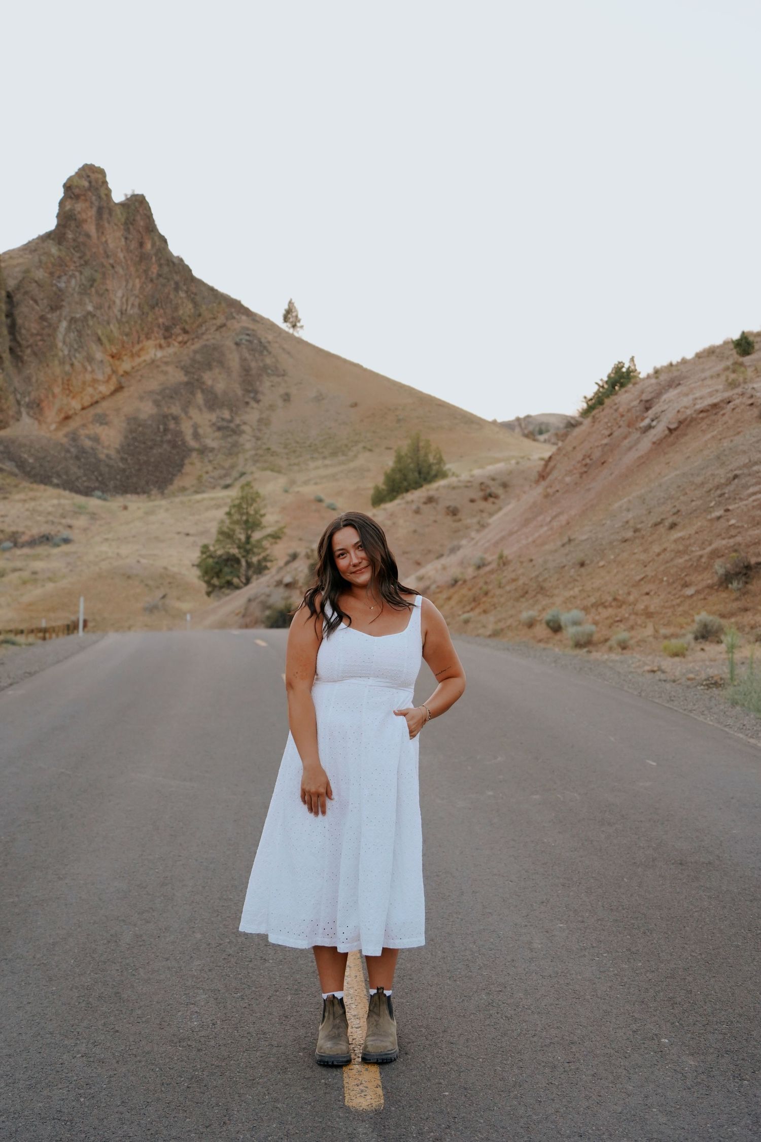 Woman in white midi dress and ankle boots standing on a scenic mountain road.