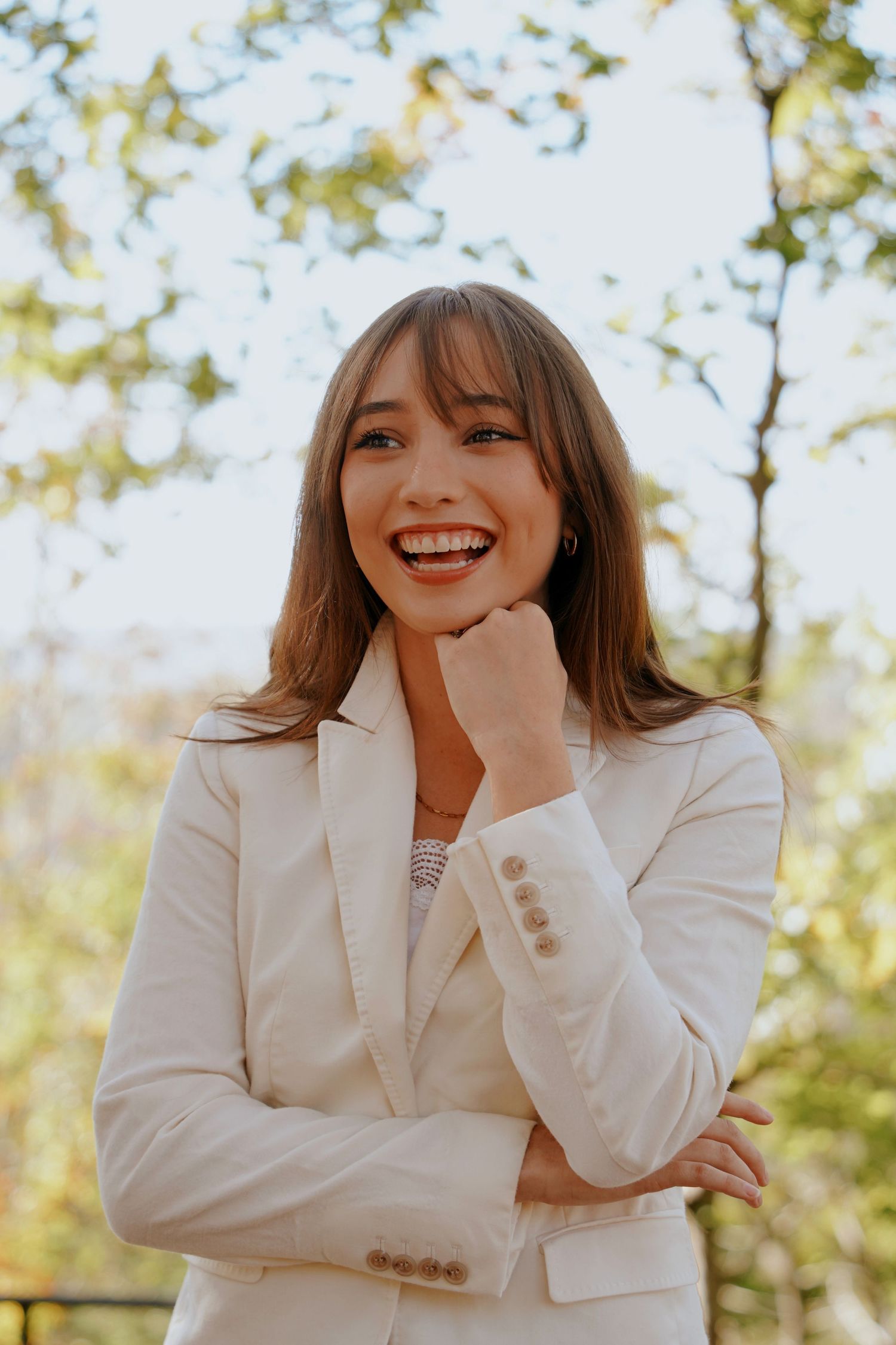 Smiling woman in white blazer posing outdoors with trees in background.
