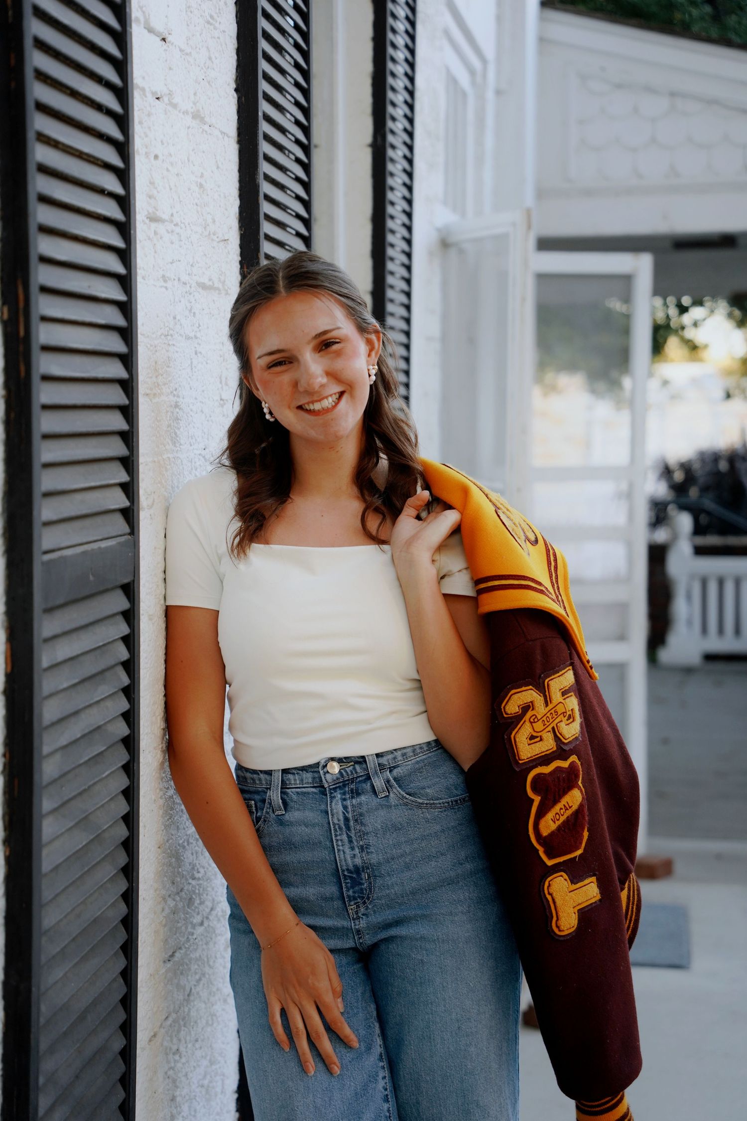 Smiling teen girl in white top holding varsity letterman jacket against white building.