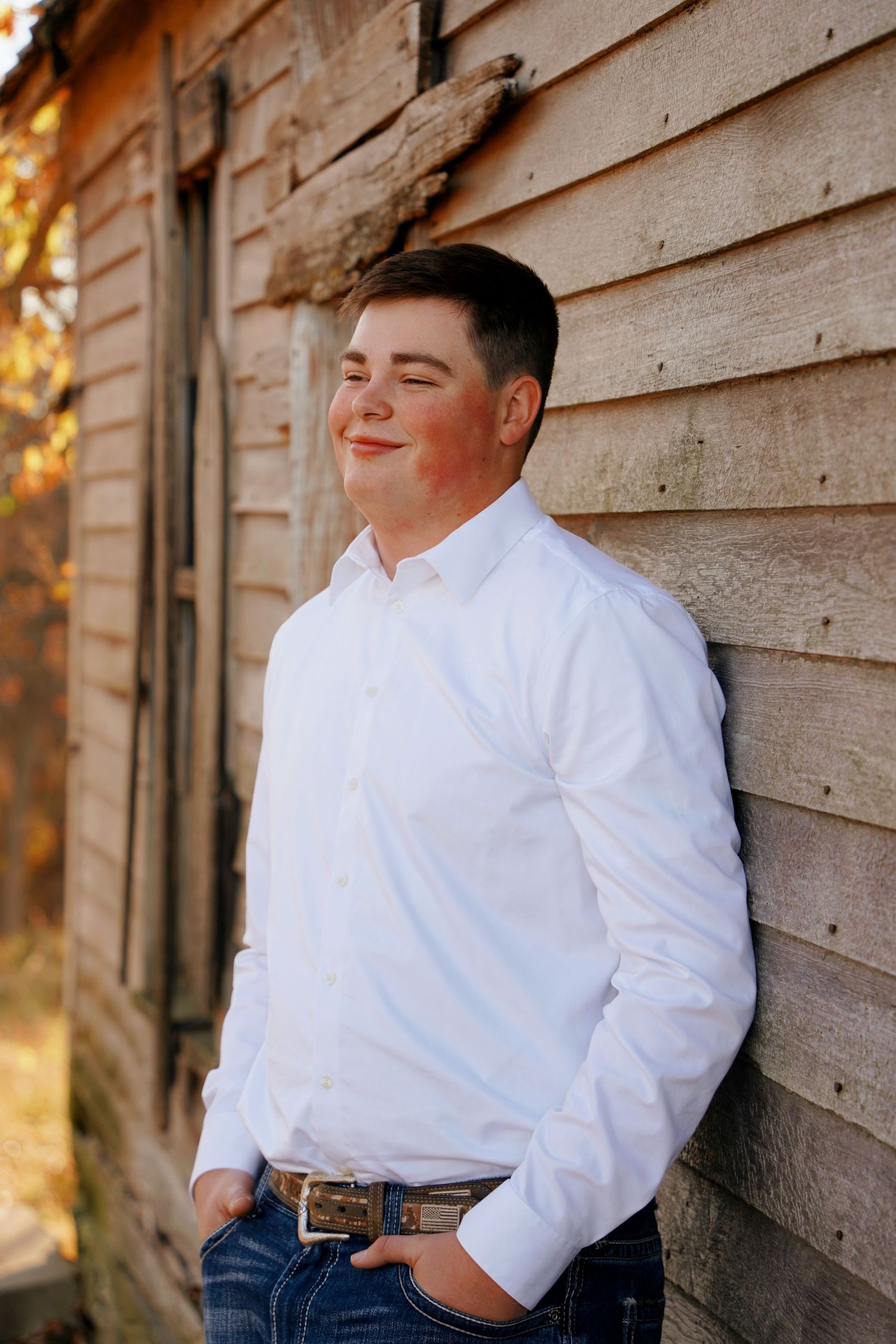 Young man in white dress shirt leaning against rustic wooden barn wall.