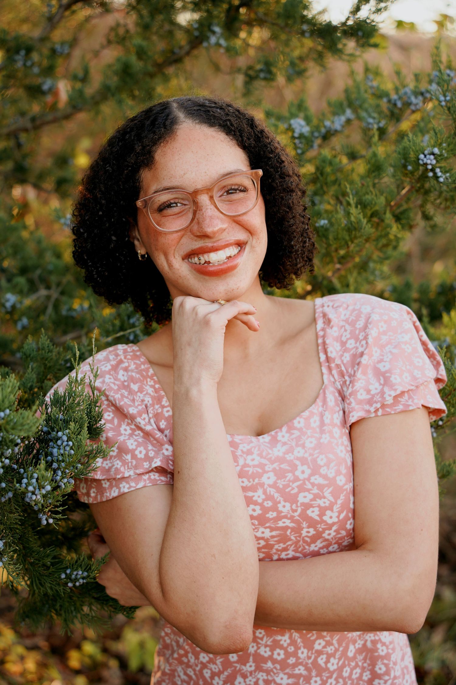 Smiling young woman with curly hair and glasses in pink floral dress outdoors.