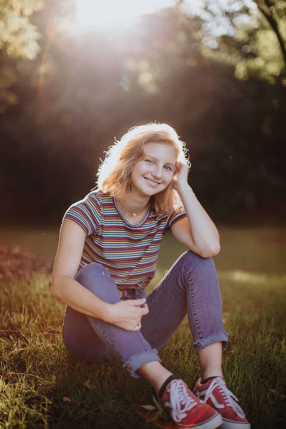 Person in striped shirt and jeans sits in sunlit grass wearing red sneakers during golden hour.