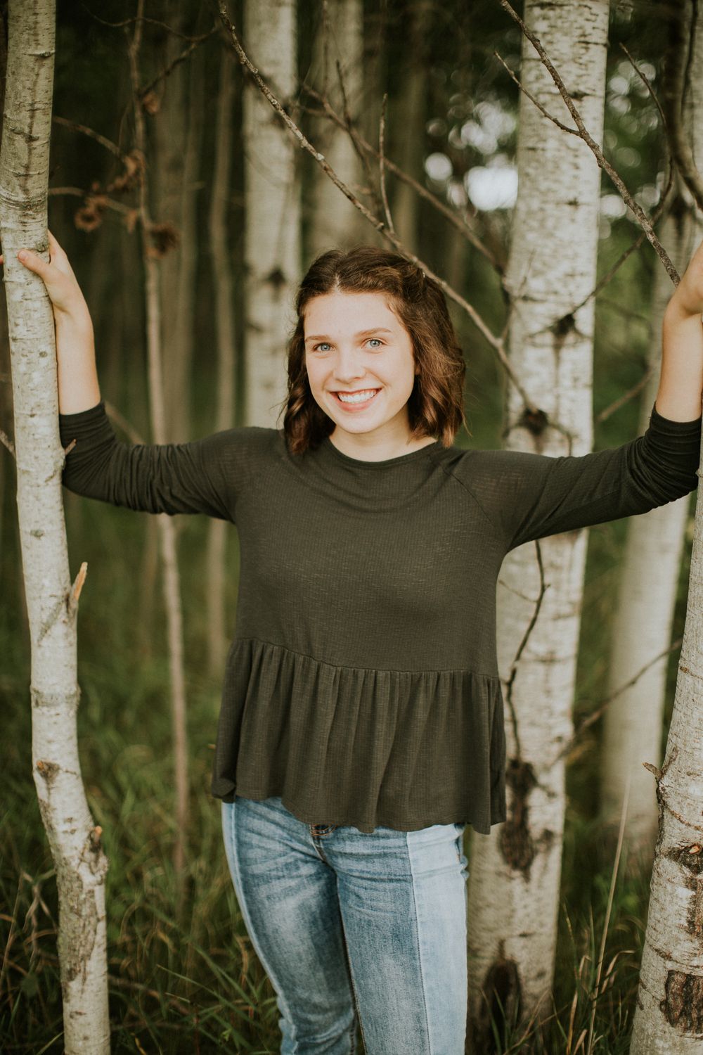 Person wearing black long sleeve top and jeans poses playfully between white birch trees in a forest setting.
