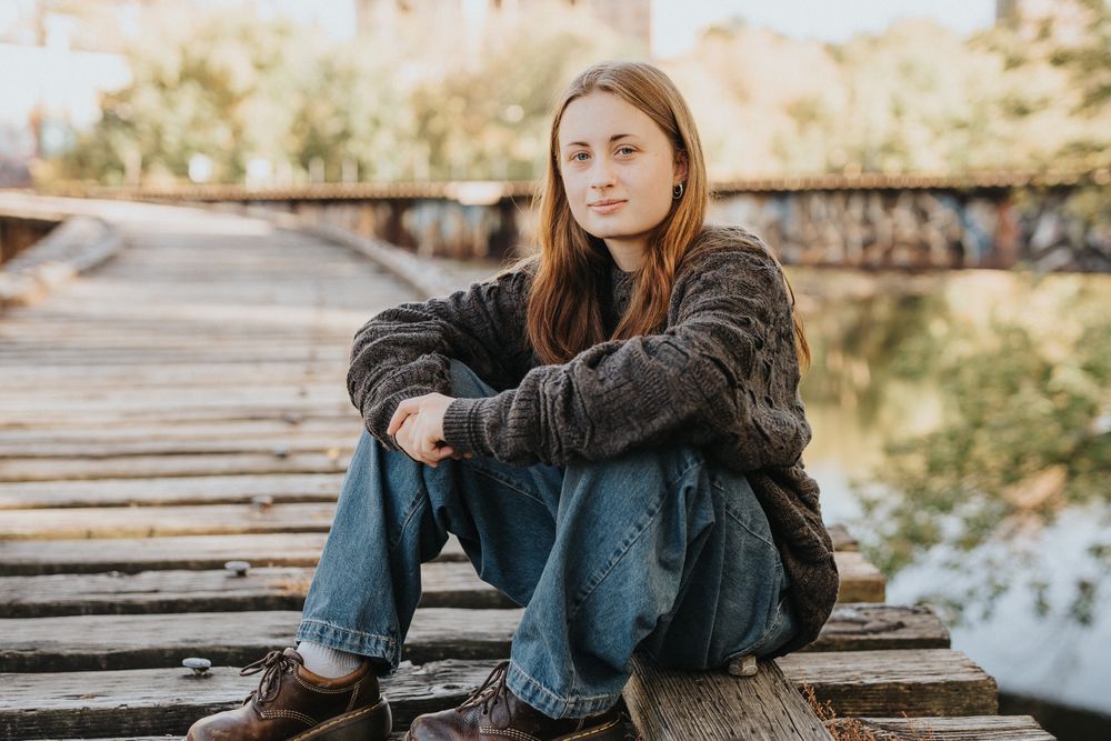 Person in casual wear sitting on wooden bridge during golden hour wearing denim jeans and dark jacket.