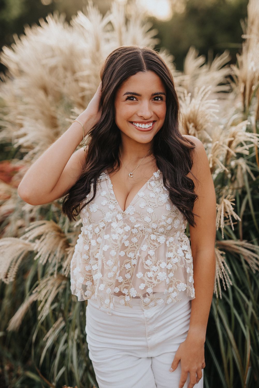 Woman in white lace dress poses outdoors against pampas grass in warm natural lighting.