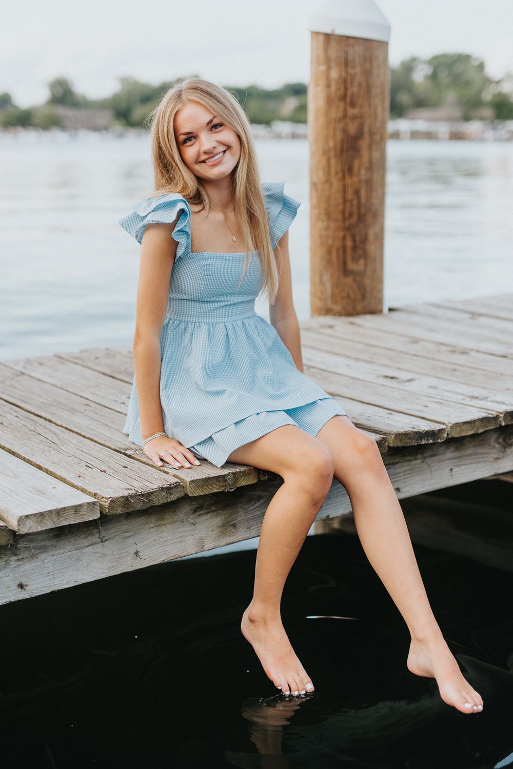 A child in a light blue dress sits on a wooden dock over water on a sunny summer day.