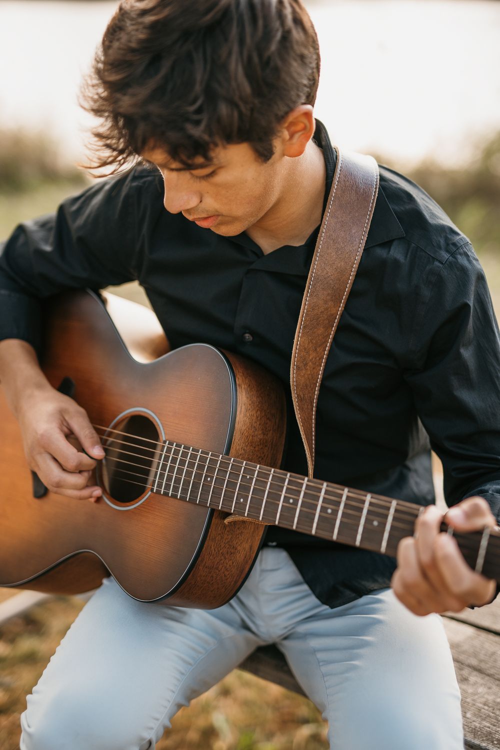 A guitarist in a black shirt and light jeans plays an acoustic guitar outdoors during golden hour.