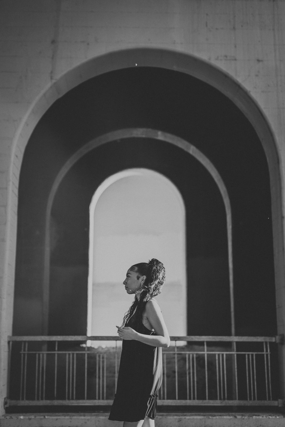 A sequence of black and white architectural arches frames a silhouetted figure leaning against a railing at sunset.