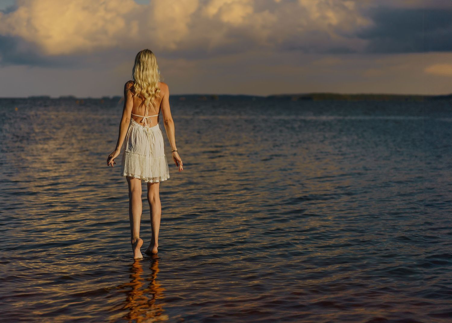 Figure in white dress standing in shallow ocean water at sunset.