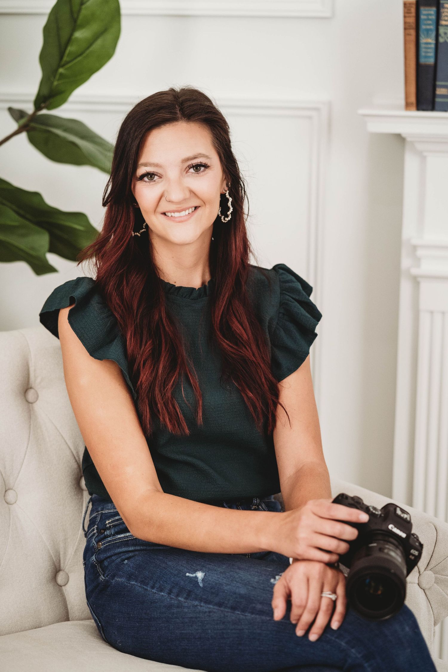 Professional photographer in a dark green top sits with camera on white armchair near indoor plant.