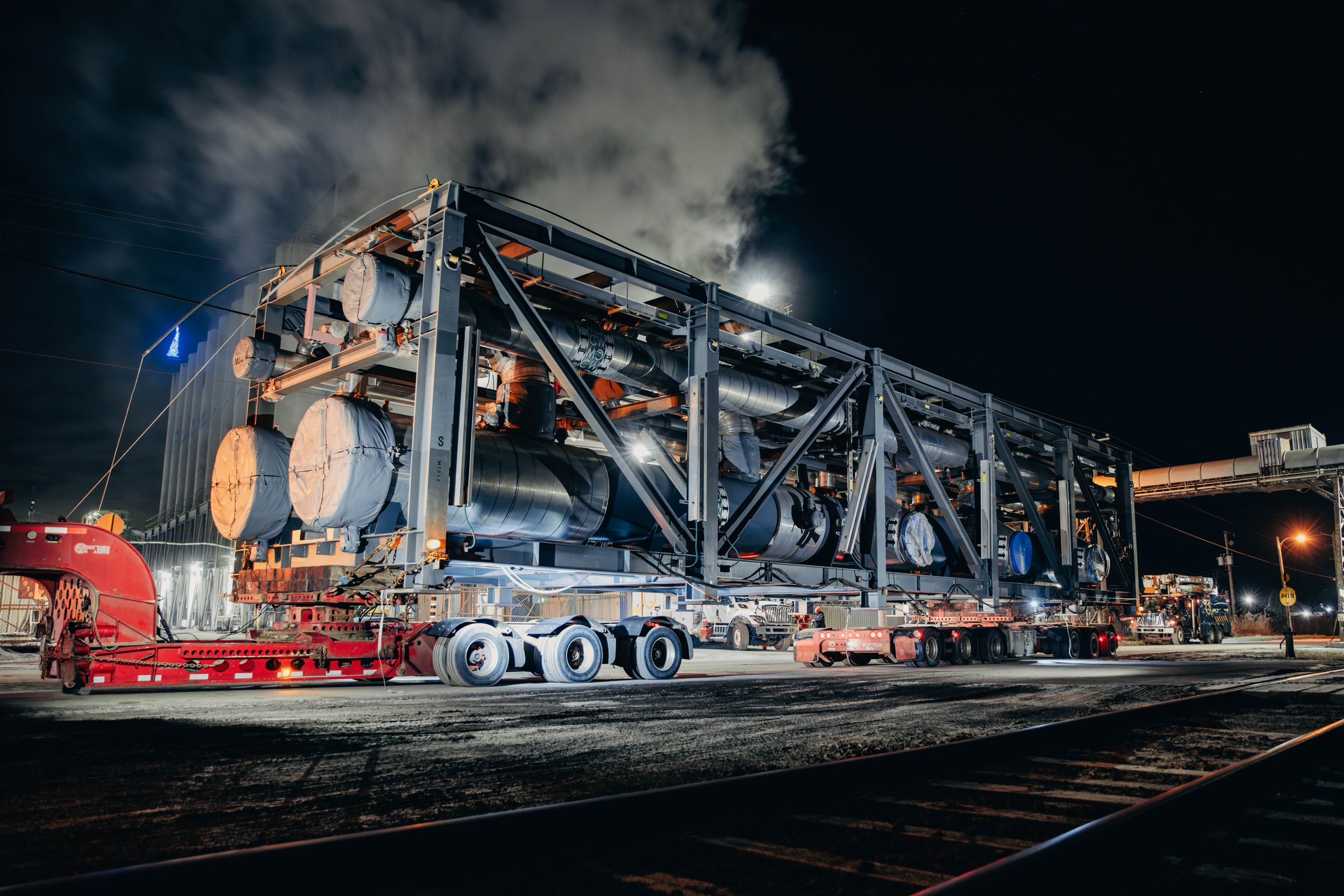 Industrial machinery being transported on a flatbed rail car at night with dramatic lighting.