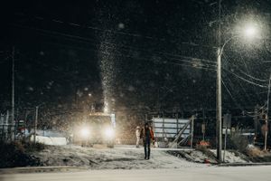 Snow falls under streetlights as figure walks through winter night scene.