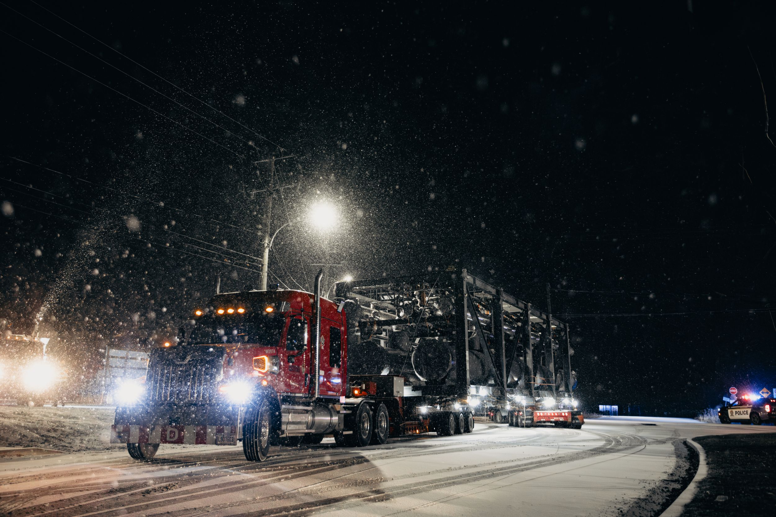 Semi truck carrying large equipment traverses snowy road at night with emergency vehicles nearby.