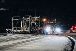 Heavy equipment and vehicles illuminate a snowy road during nighttime construction work.