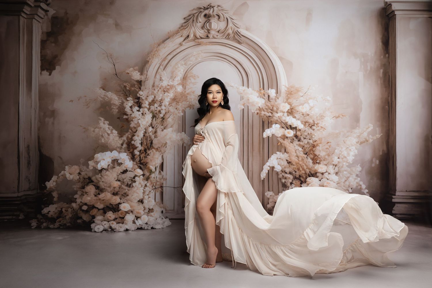 Elegant maternity photoshoot in front of ornate archway with flowing white gown and floral decorations.