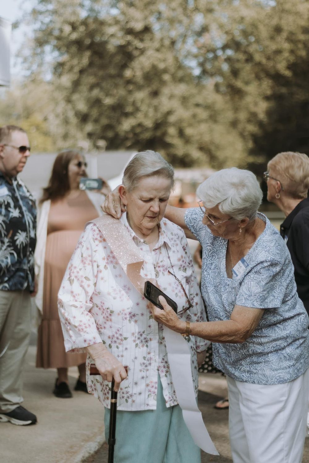 Two elderly women in floral and gray outfits enjoy a sweet moment dancing together outdoors in soft natural lighting.