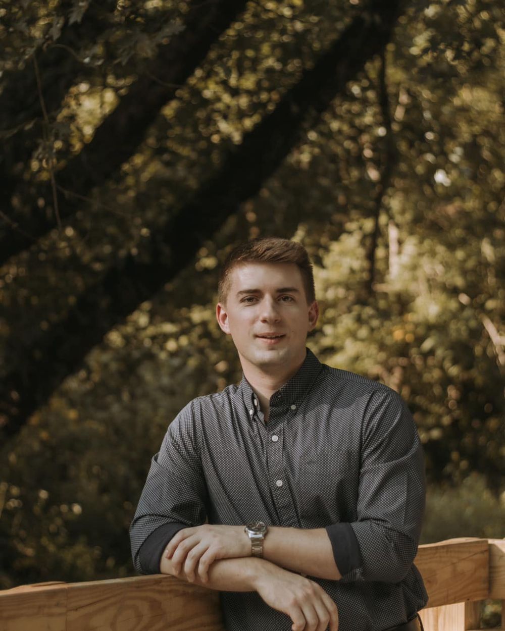 Someone in a gray button-up shirt sits in an outdoor setting with blurred trees in the background during golden hour.