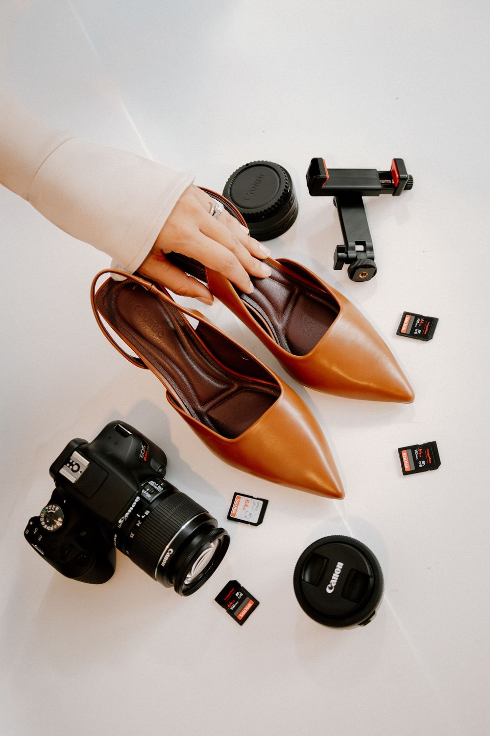 Brown leather stiletto heels arranged with professional camera equipment and memory cards on white background.