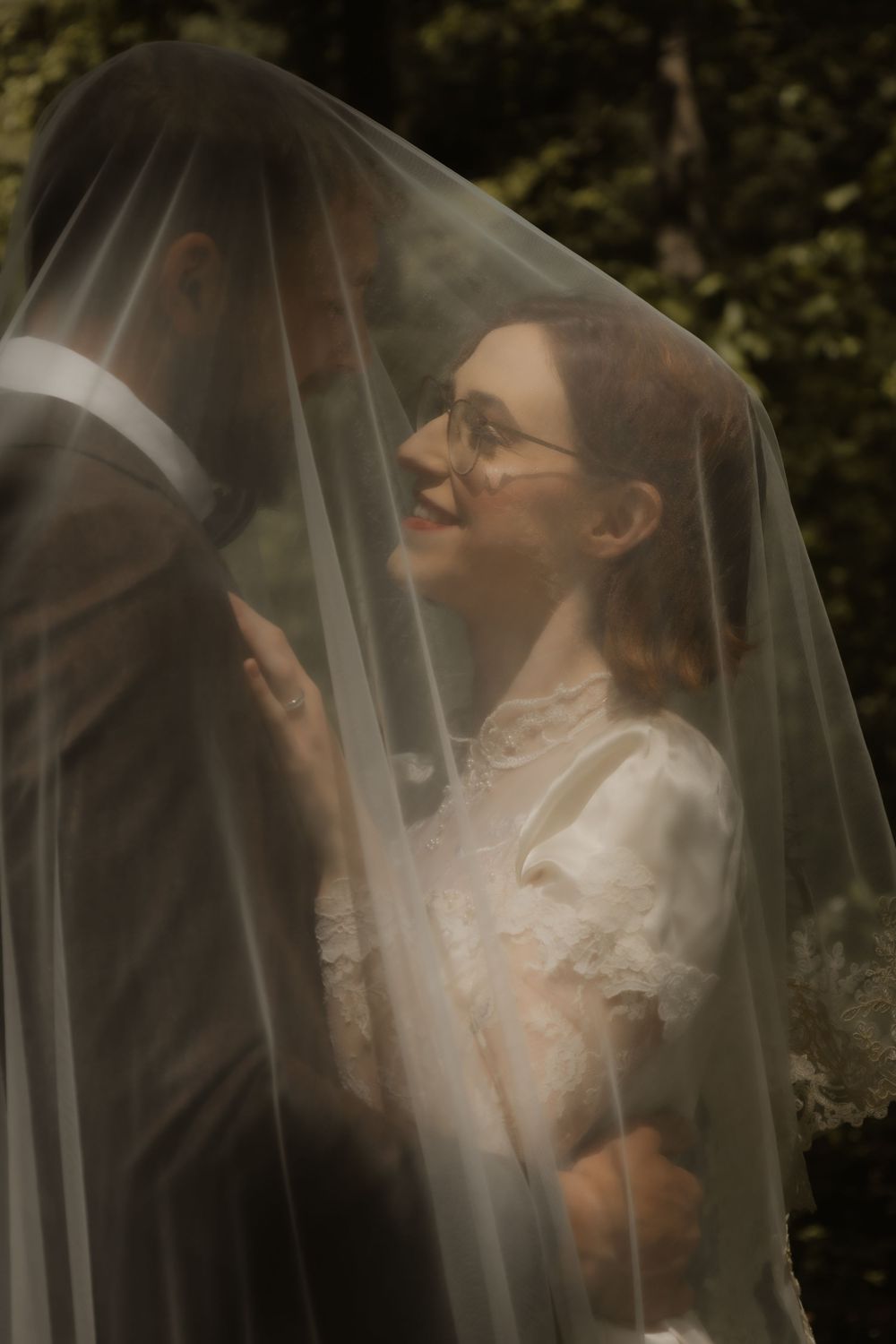 Dreamy wedding scene with couple behind flowing veil in dappled forest light.
