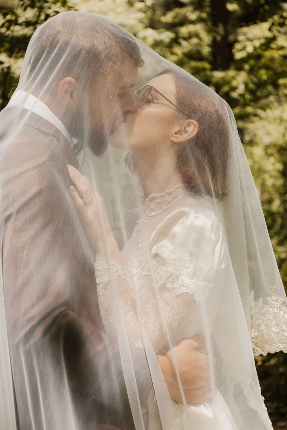 Wedding couple embraces under delicate veil with forest scenery behind them.