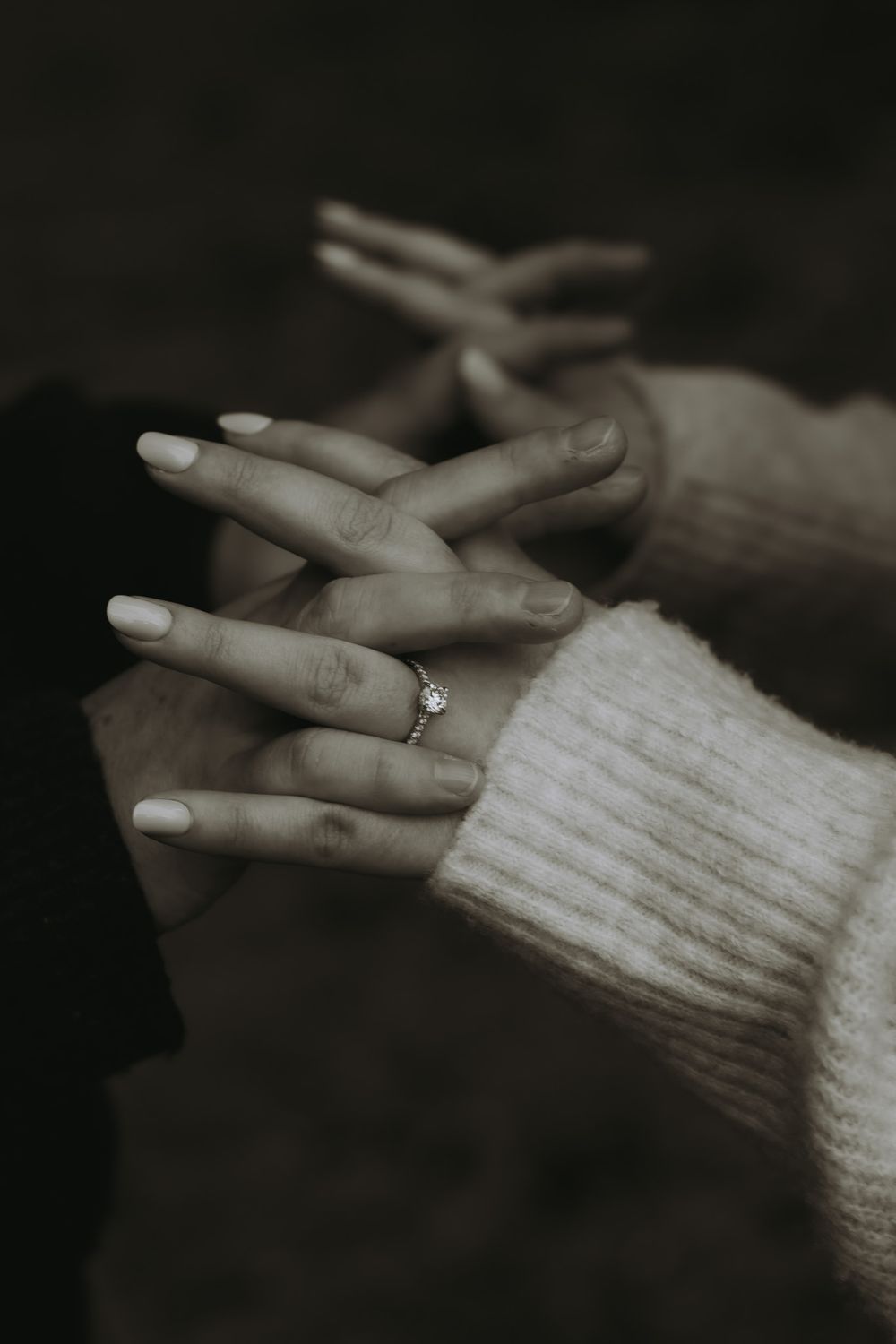 A hand wearing a knitted sweater sleeve displays an engagement ring against a dark background.