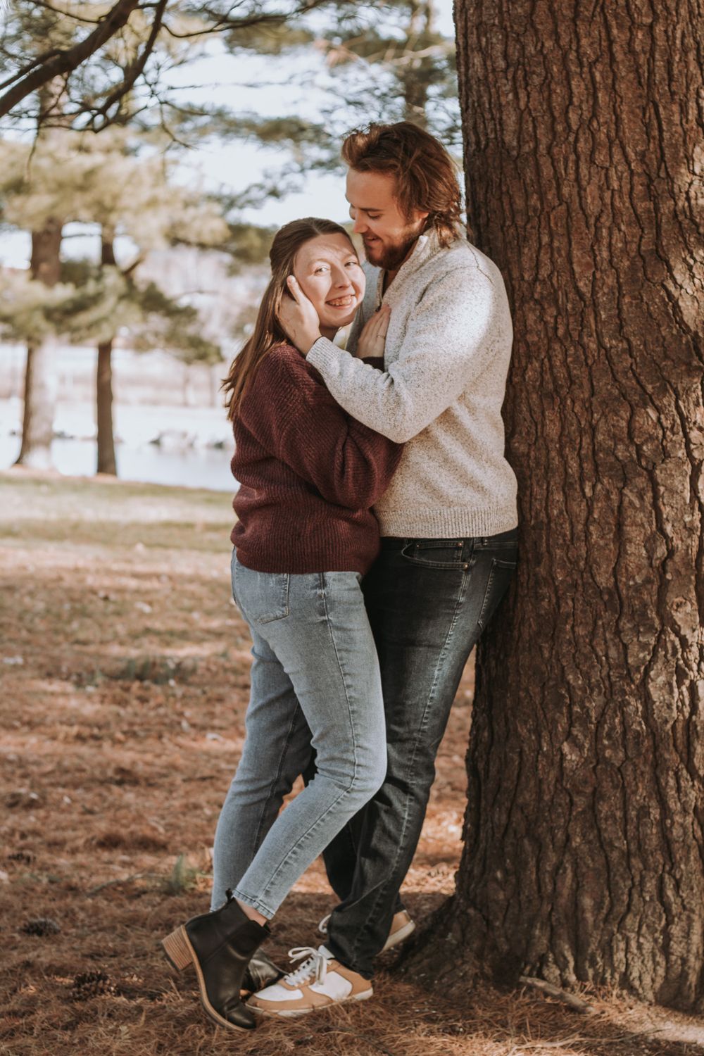 A romantic couple stands intimately near a large tree trunk in a park during an autumn afternoon.