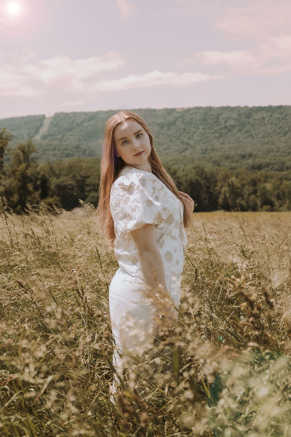 Ethereal portrait in a sunlit meadow showcases a flowing white lace dress against rolling hills.