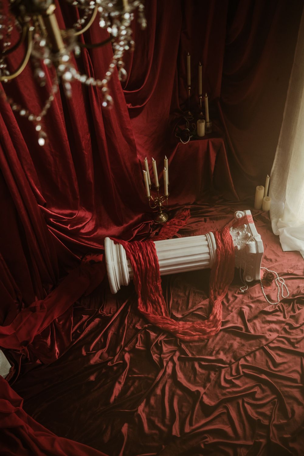 A red fabric set with candles, a chandelier hanging above, and a column laying on the floor.