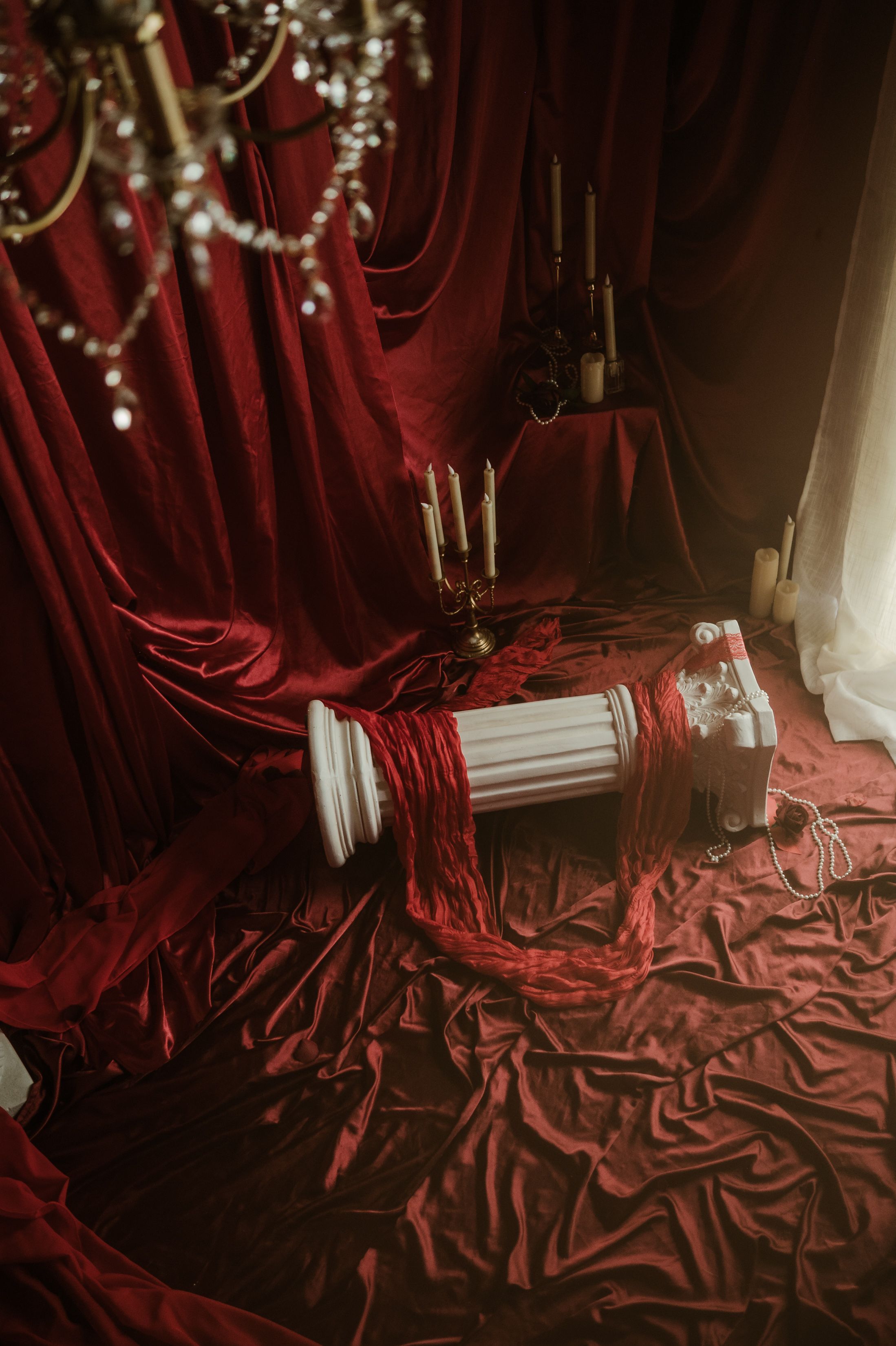 A red fabric set with candles, a chandelier hanging above, and a column laying on the floor.