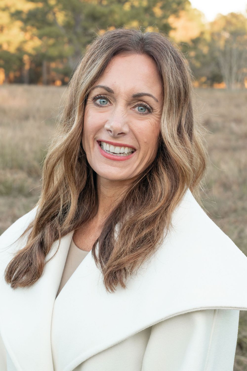 A woman in a white coat smiles warmly at the camera during an outdoor photoshoot.