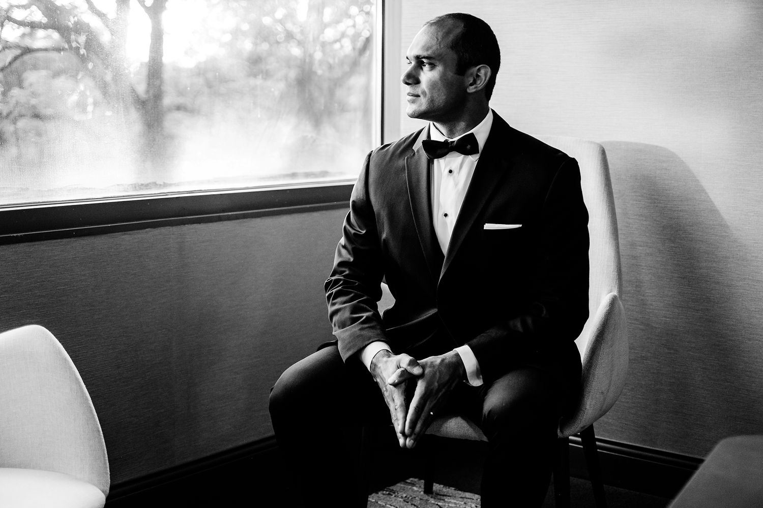 Classic black and white portrait of groom in tuxedo seated by window.