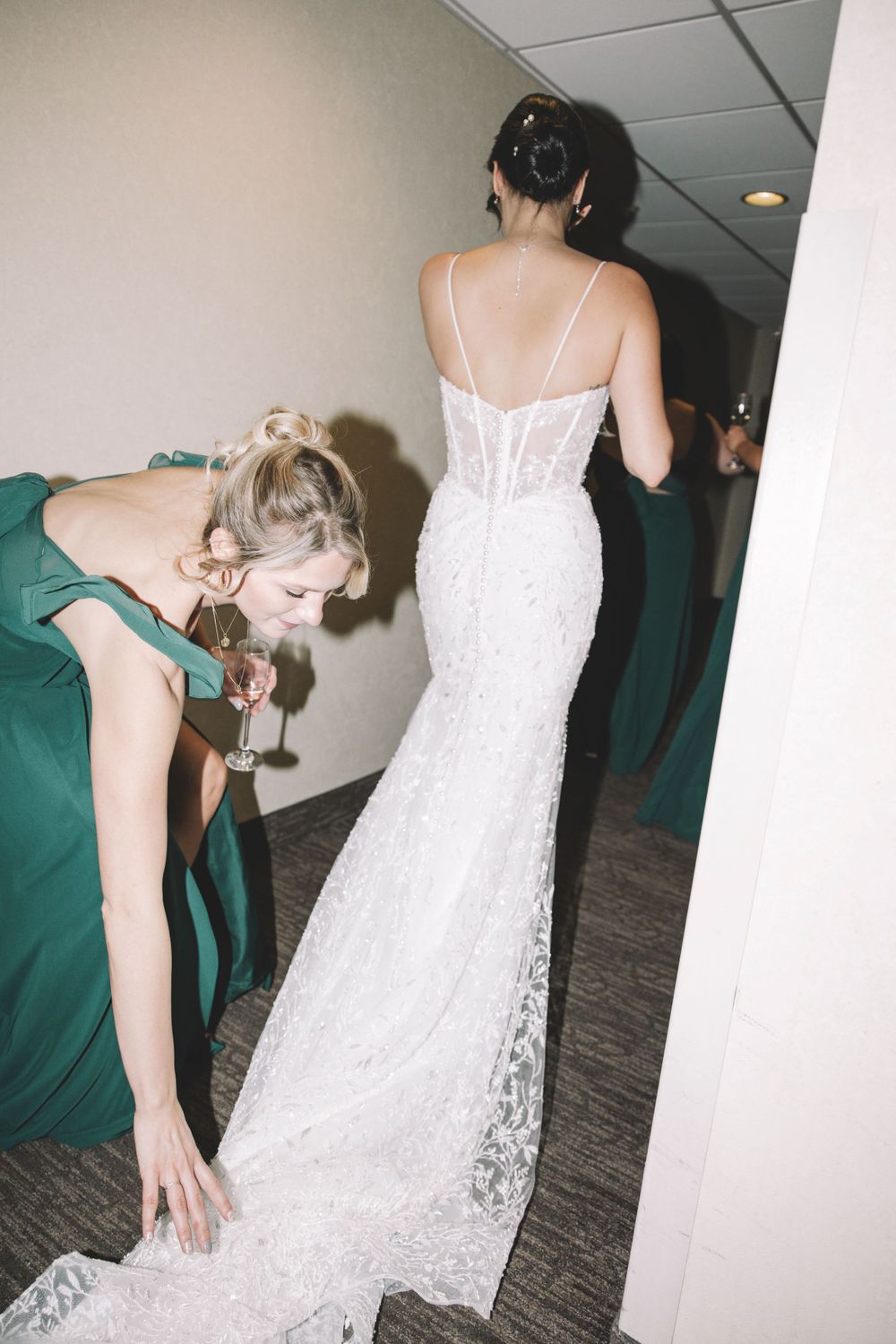 Bridesmaids help arrange the train of a lace wedding dress in a hallway.