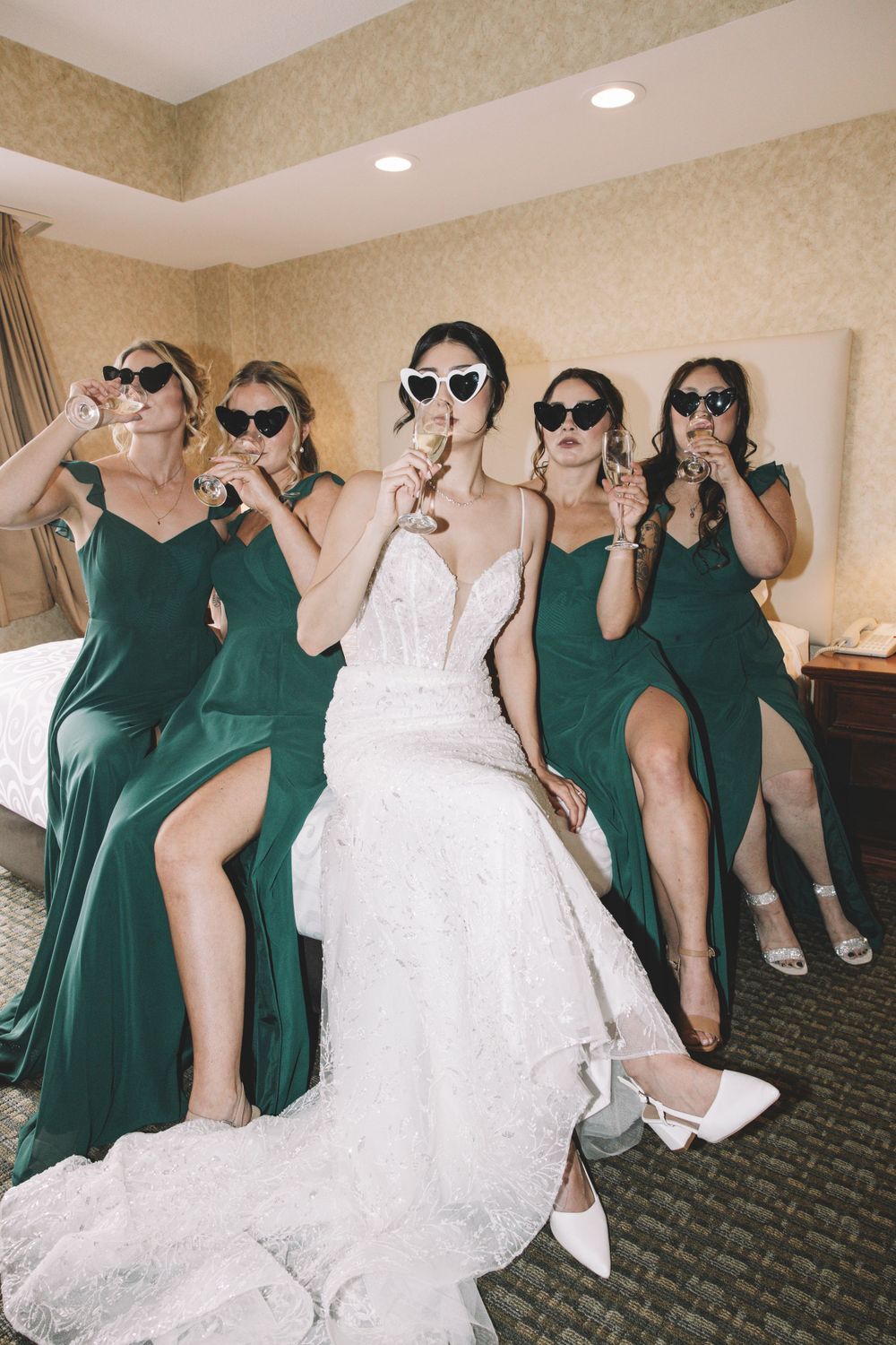 Group wearing sunglasses pose playfully in formal dresses at wedding celebration.