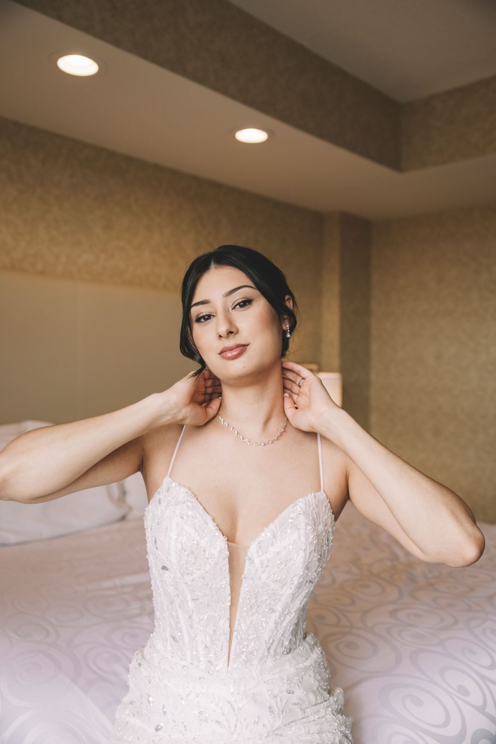 A bride wearing an elegant white beaded wedding gown with plunging neckline poses in a room with recessed lighting.