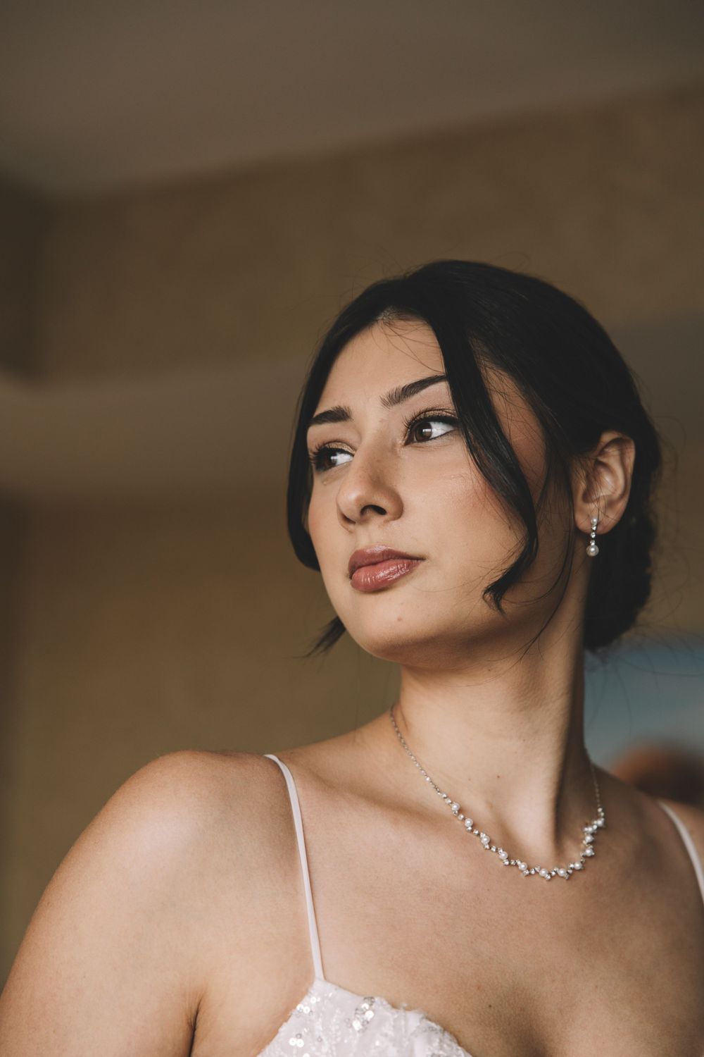 A portrait of someone wearing a white dress with spaghetti straps and sparkling necklace looking to the side.