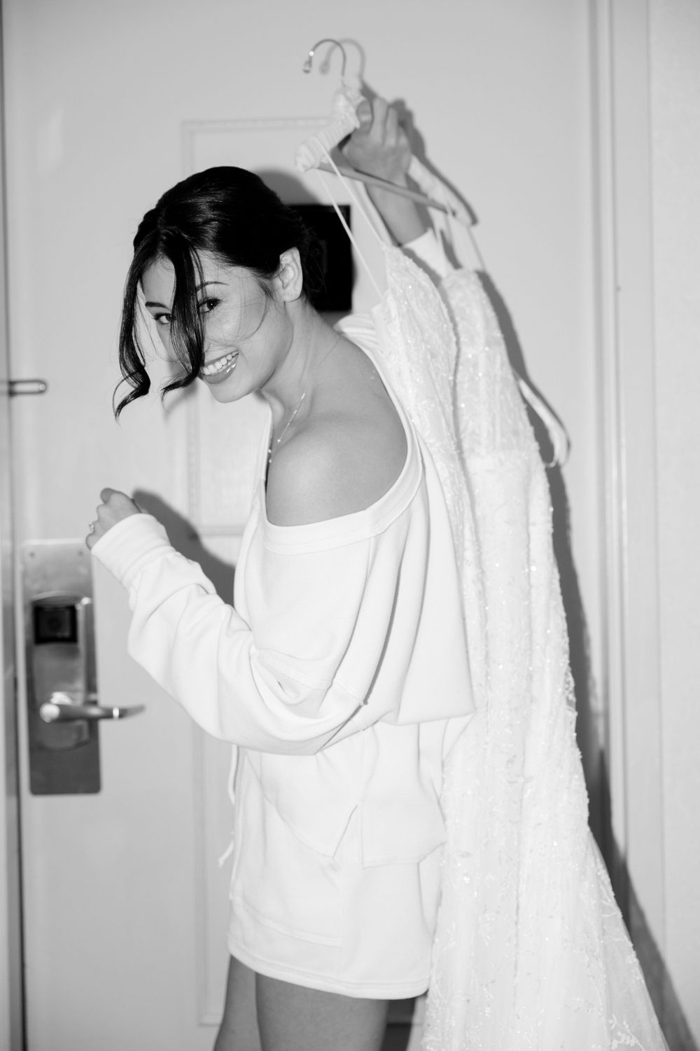 Black and white photo of person in white robe laughing while white wedding dress hangs in background.