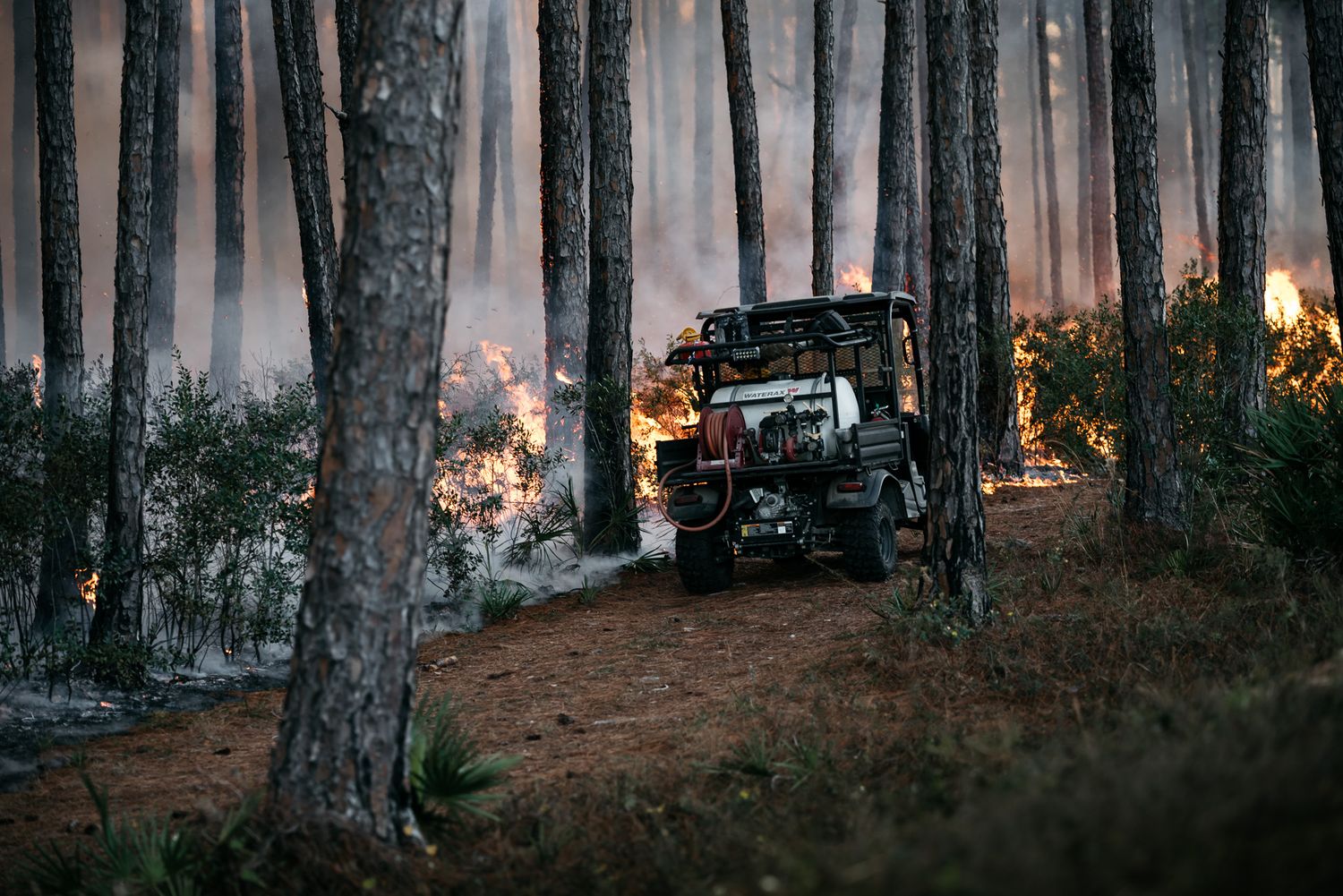Off-road vehicle traverses through smoky forest with wildfire burning in background.