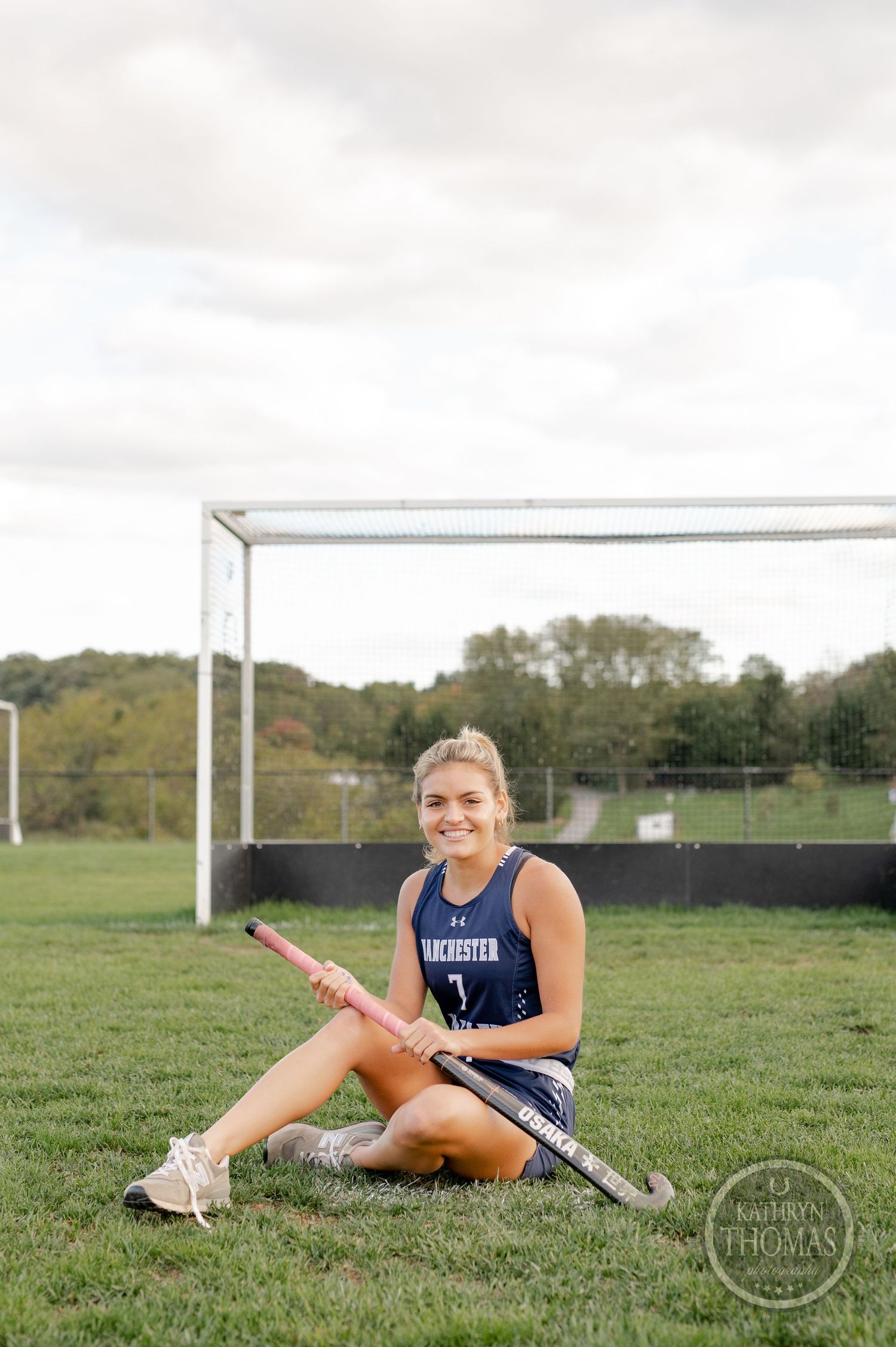 Field hockey player sits on grass field with stick by goal post in navy blue uniform.