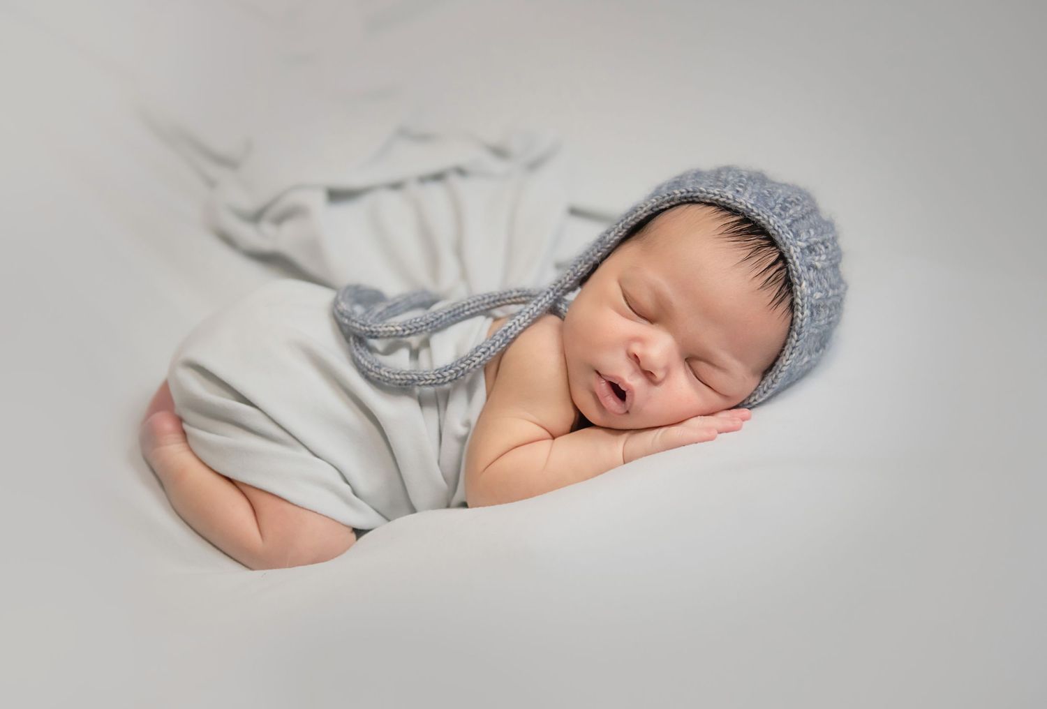 Sleeping newborn wearing grey knit bonnet on soft white background.