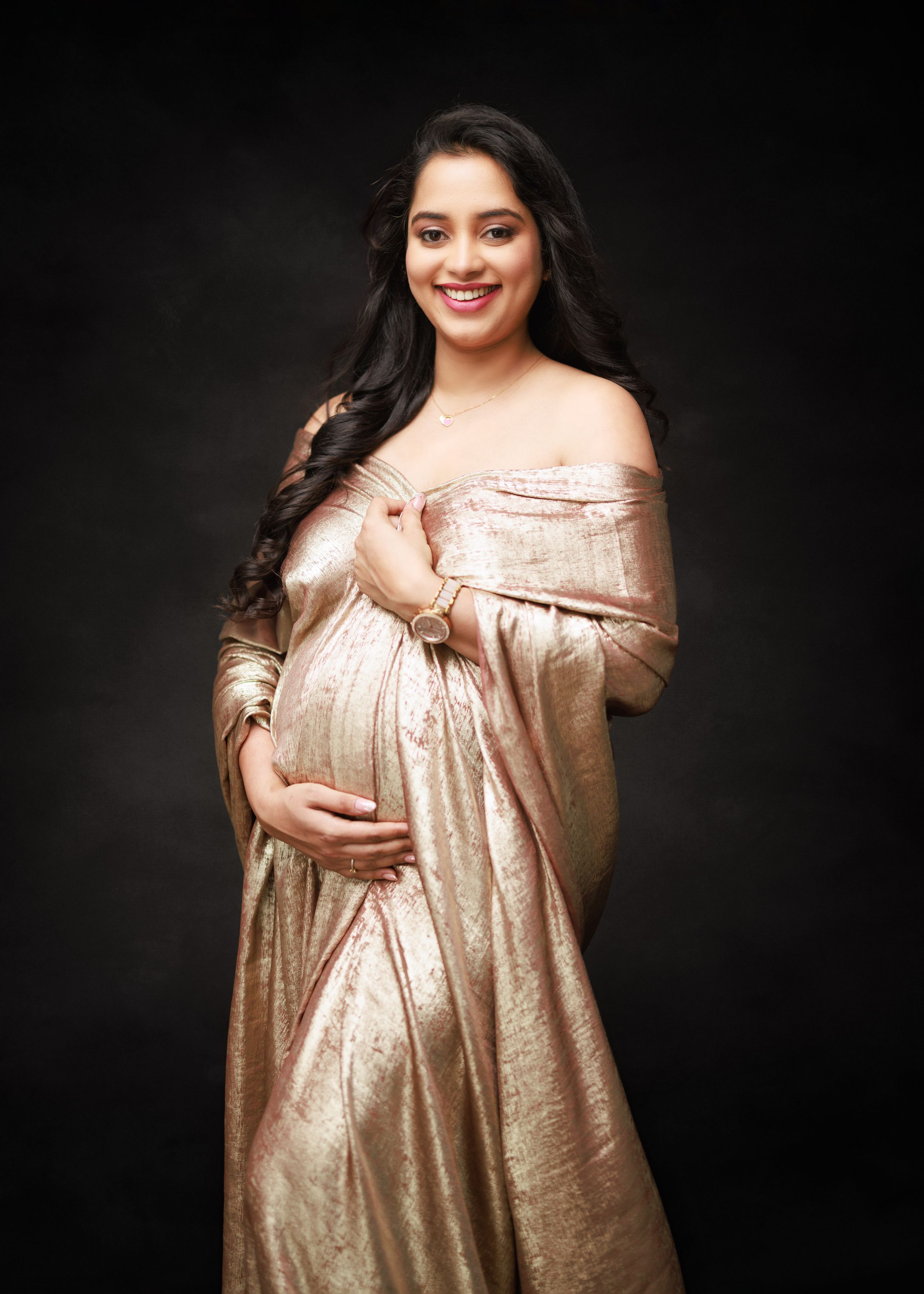 A glamorous maternity portrait featuring a champagne-colored off-shoulder gown against dark background.