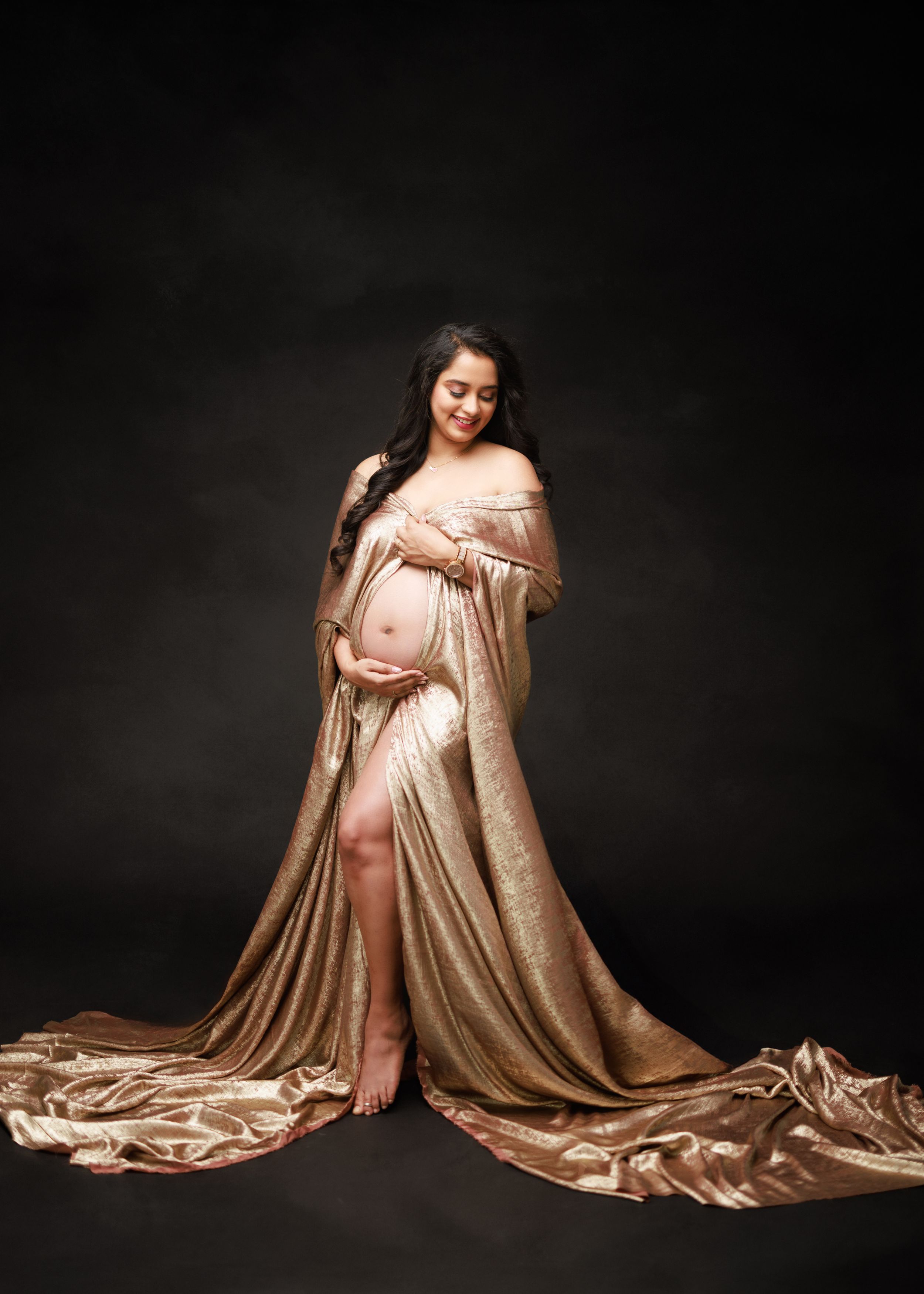 A dramatic maternity photo with flowing champagne silk gown against dark backdrop.