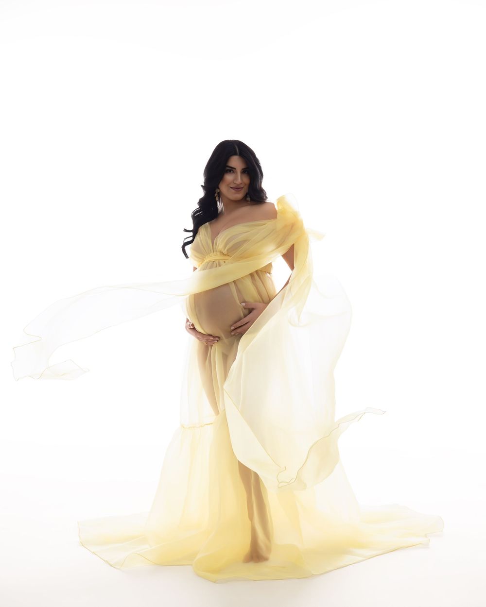 Pregnant woman in flowing yellow tulle maternity gown poses gracefully against white background in professional photo shoot.