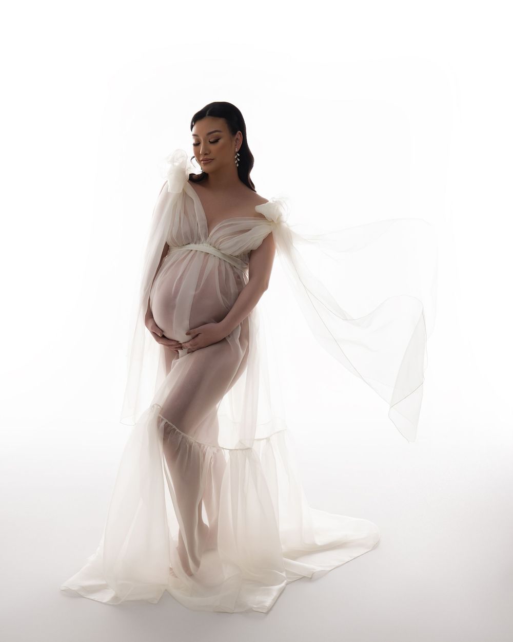 An ethereal maternity photo featuring a flowing sheer white gown against a light background in a dreamy studio setting.